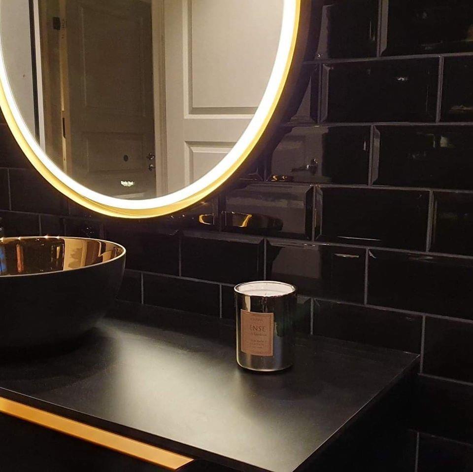 A round lit mirror over a dark vanity in black tiles